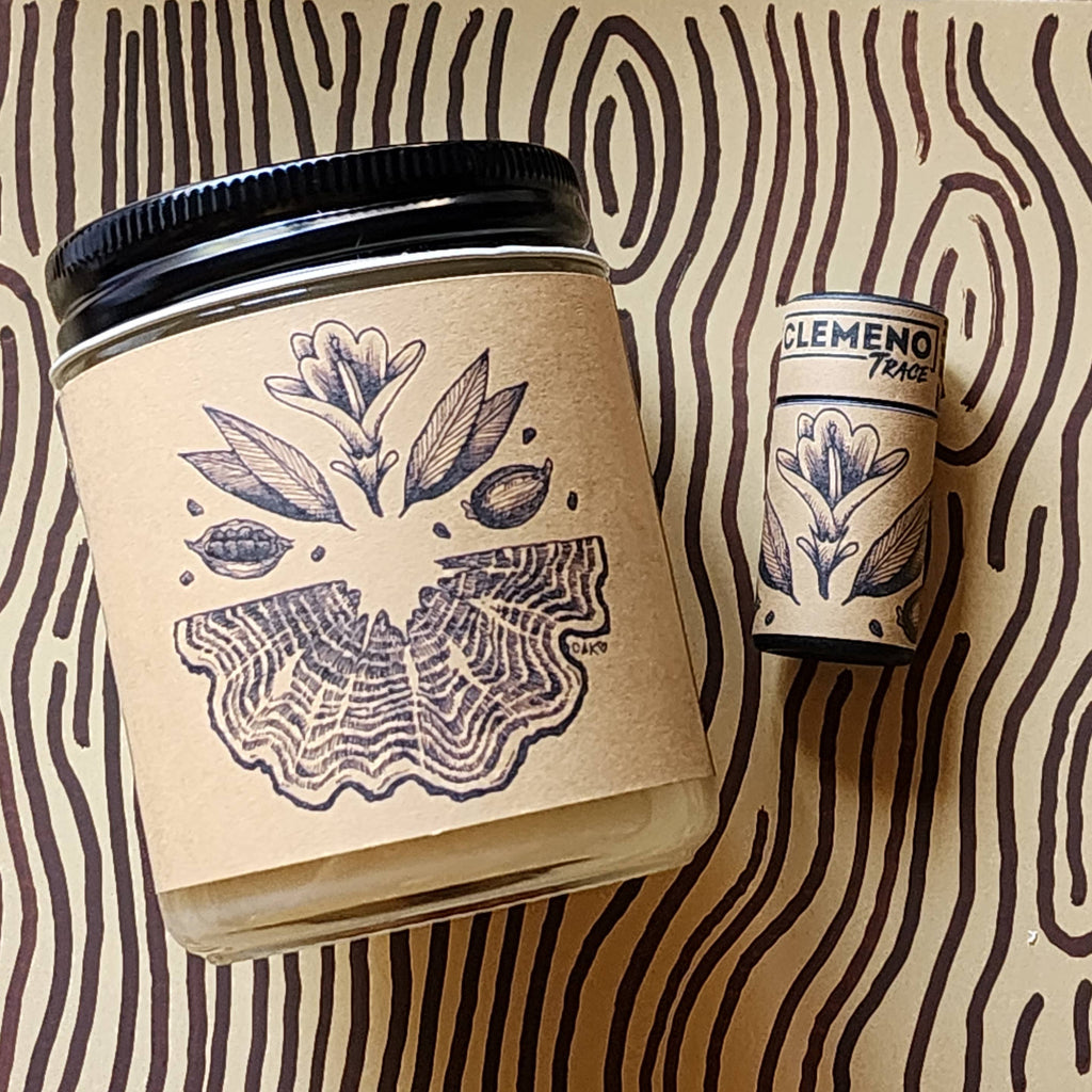 A clear glass jar candle with a black lid and a kraft paper label featuring a botanical illustration, shown next to a matching cylindrical tin on a beige background with dark brown wavy lines.