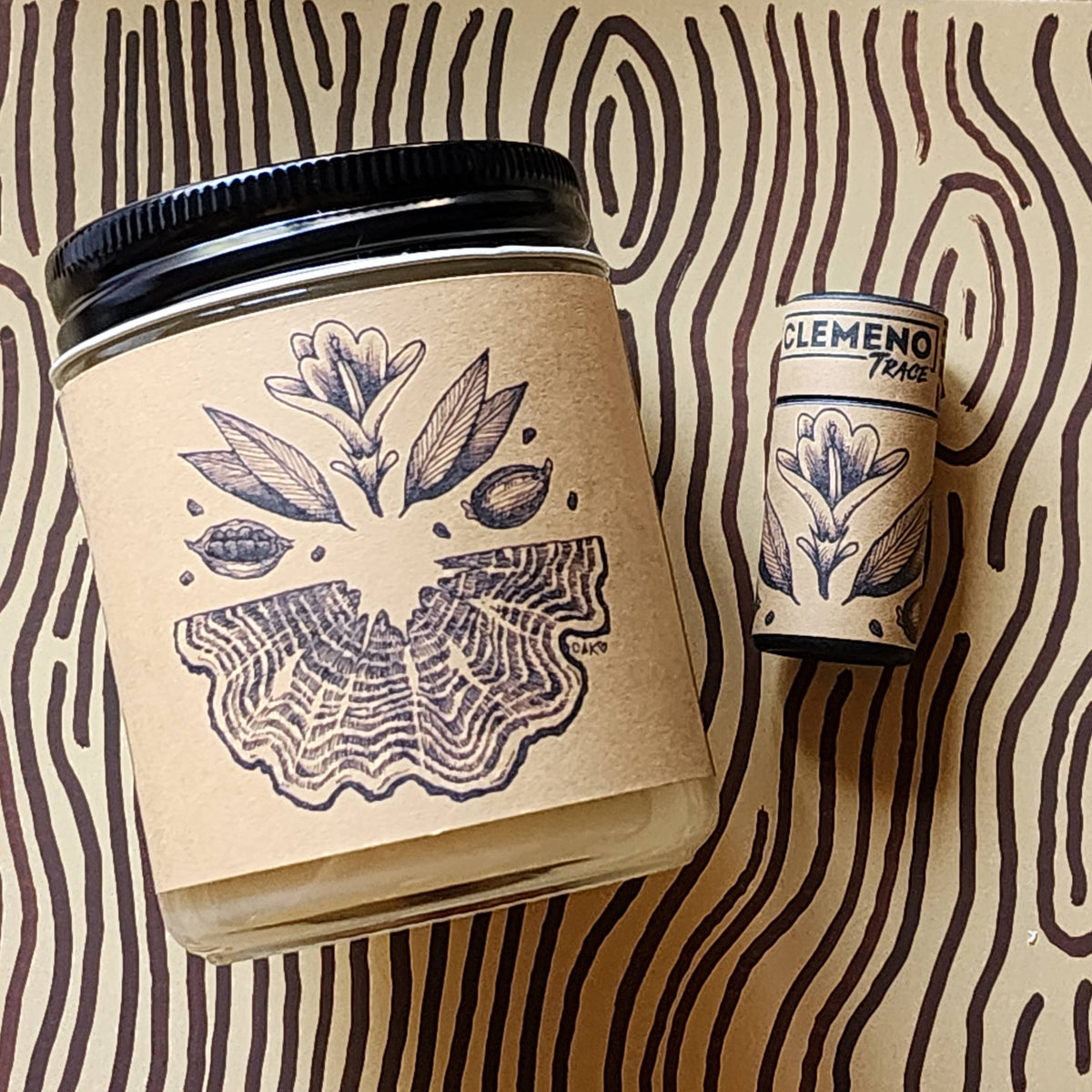 A clear glass jar candle with a black lid and a kraft paper label featuring a botanical illustration, shown next to a matching cylindrical tin on a beige background with dark brown wavy lines.