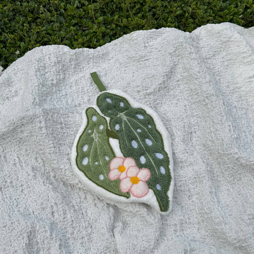 A handcrafted hand towel shaped like a green leaf with white spots and a pink flower with a yellow center, resting on a textured white blanket with green foliage in the background.