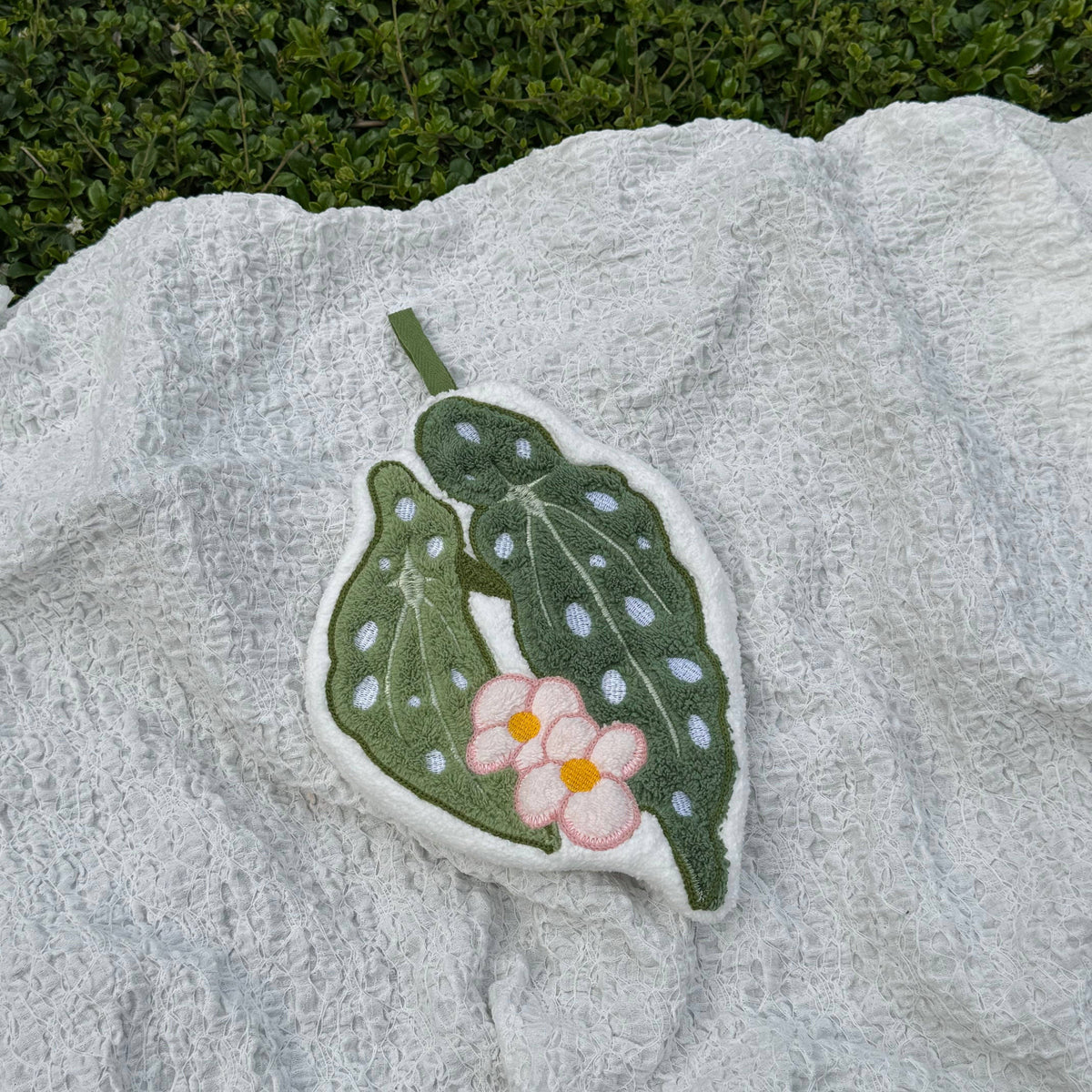 A handcrafted hand towel shaped like a green leaf with white spots and a pink flower with a yellow center, resting on a textured white blanket with green foliage in the background.