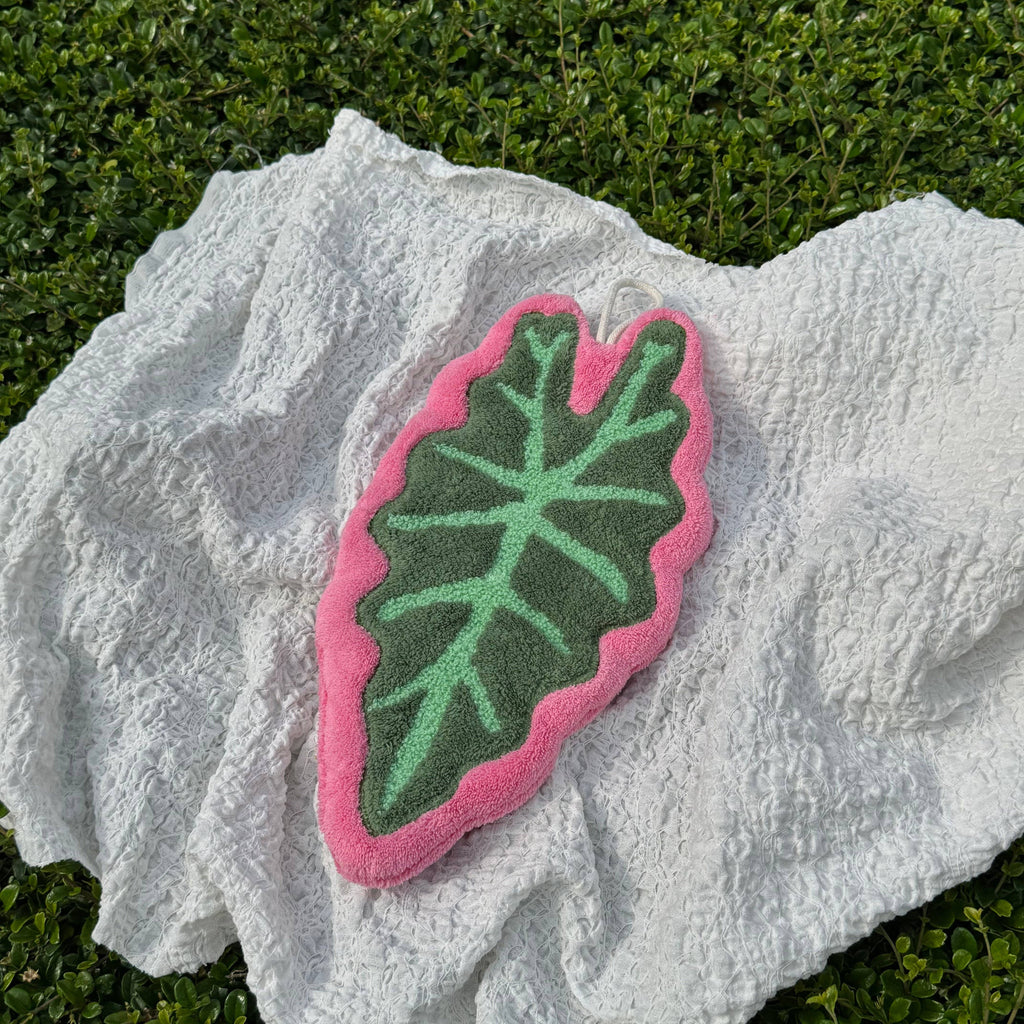 A leaf-shaped hand towel with a pink border and green leaf vein pattern, resting on a white textured towel over green grass.