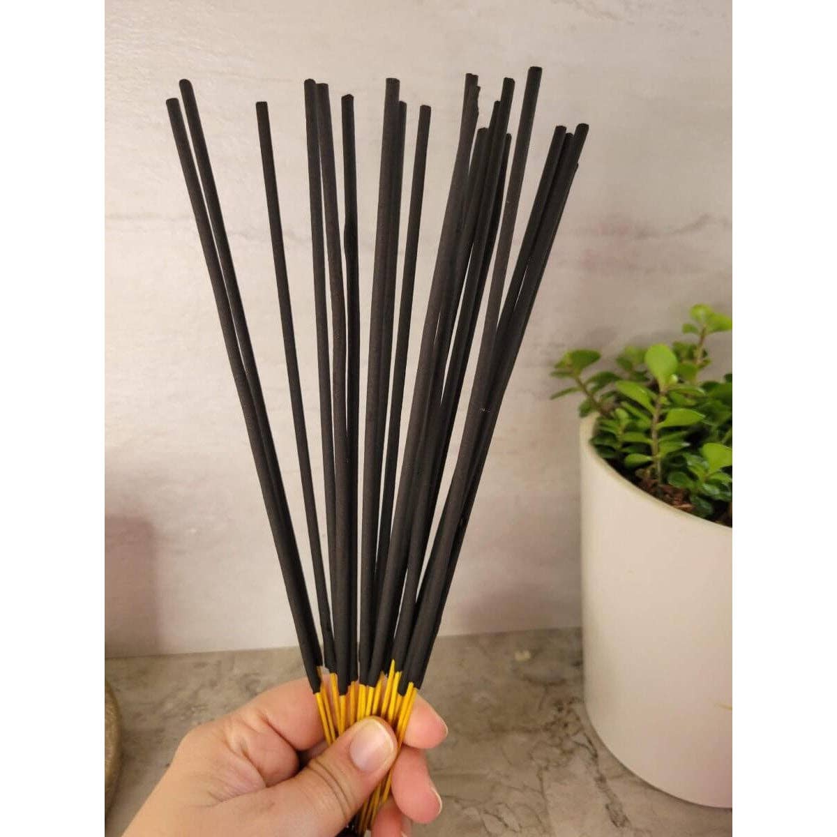 Hand holding a bundle of black incense sticks with gold tips, against a neutral background.