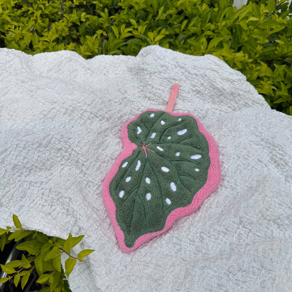 A handcrafted leaf-shaped hand towel with a dark green center, white spots, and a pink border, placed on a textured white blanket with green foliage in the background.