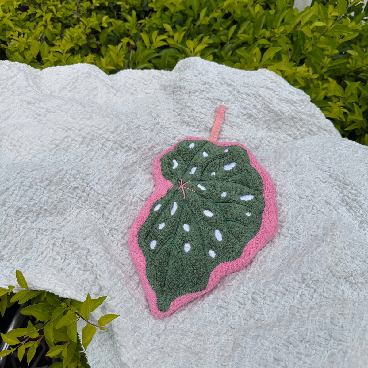 A handcrafted leaf-shaped hand towel with a dark green center, white spots, and a pink border, placed on a textured white blanket with green foliage in the background.