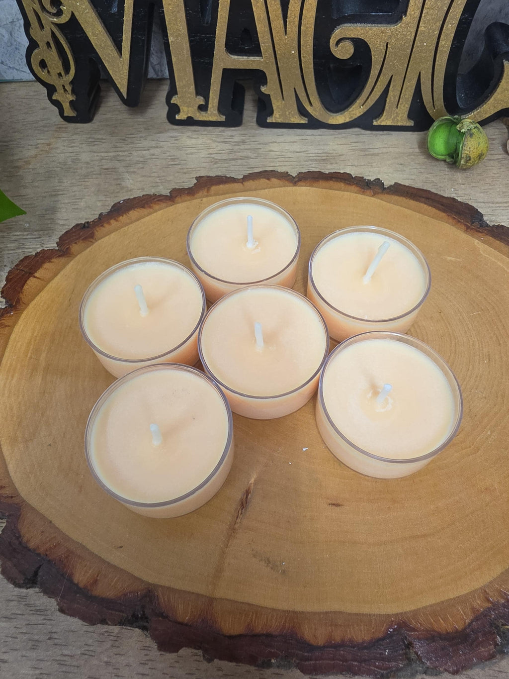 Five small beige tea light candles arranged on a round natural wood slice tray, with a decorative gold and black sign in the background.