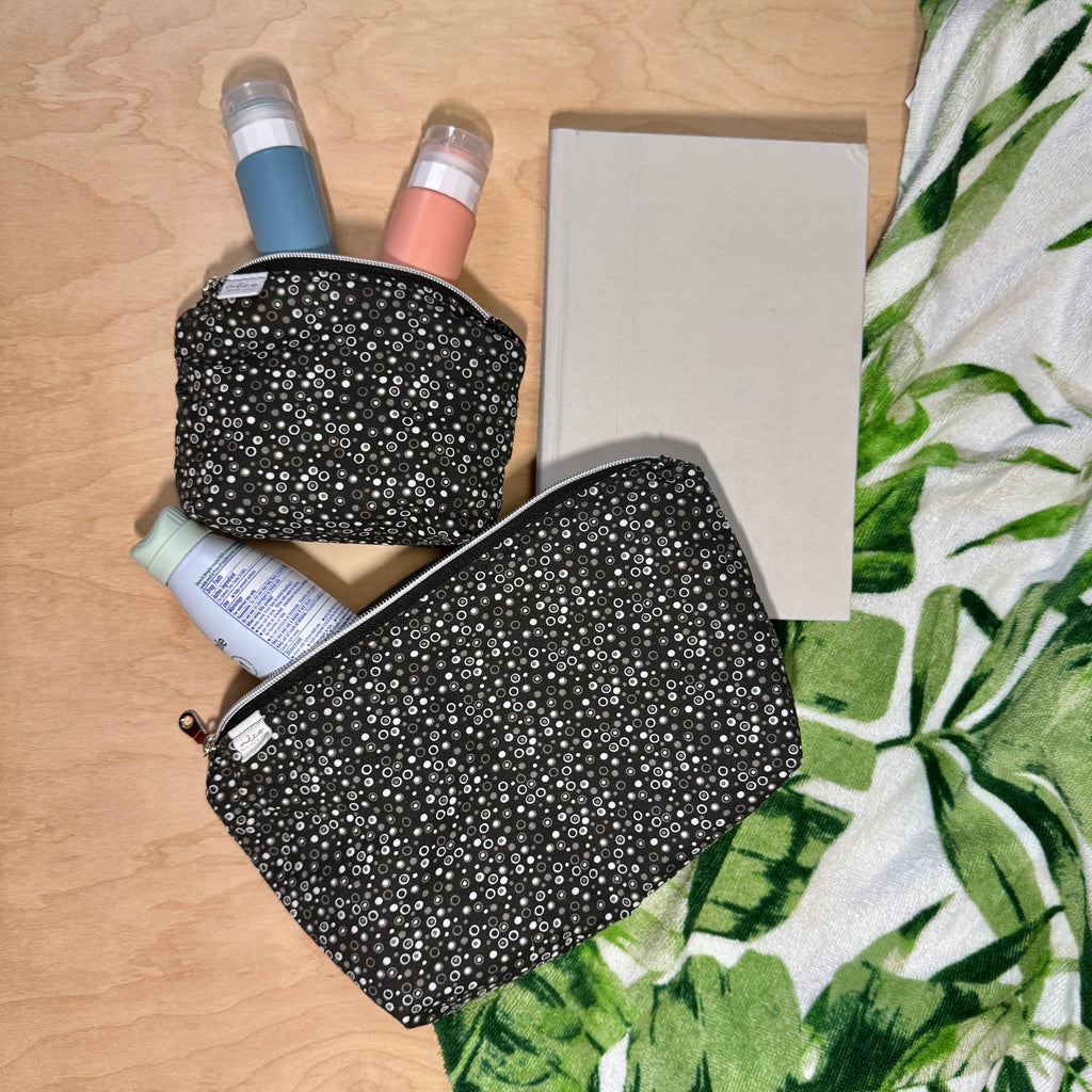 Two black zippered cosmetic with a small white floral print are arranged on a light wood surface alongside a notebook and travel-sized bottles. A green-and-white tropical leaf patterned fabric is visible on the right side.