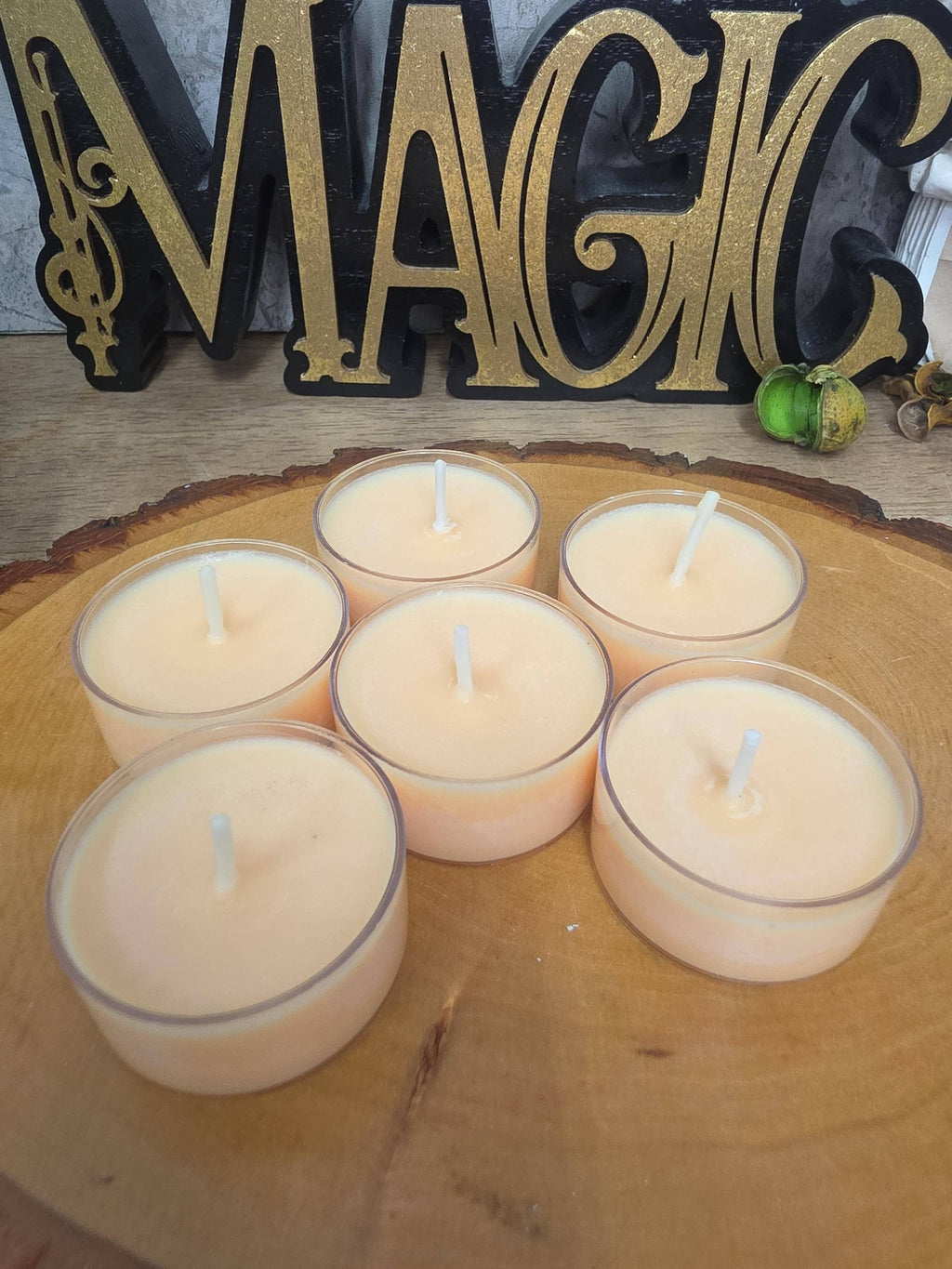 Five small tea light candles with white wicks in clear glass holders, arranged on a round wooden slice board. A decorative gold and black 'MAGIC' sign is visible in the background.