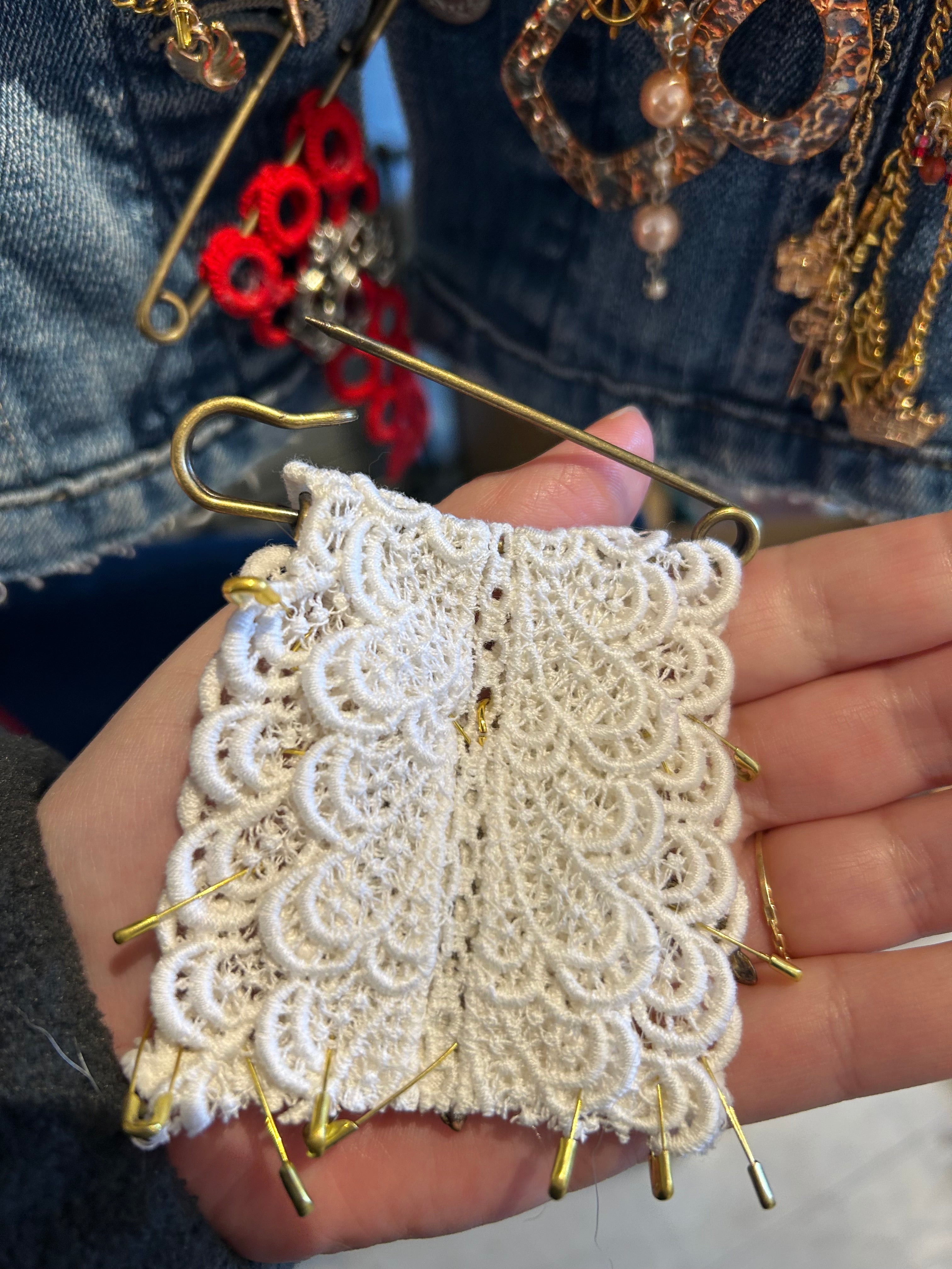 Hand holding a piece of lacework with pins, surrounded by other jewelry items.