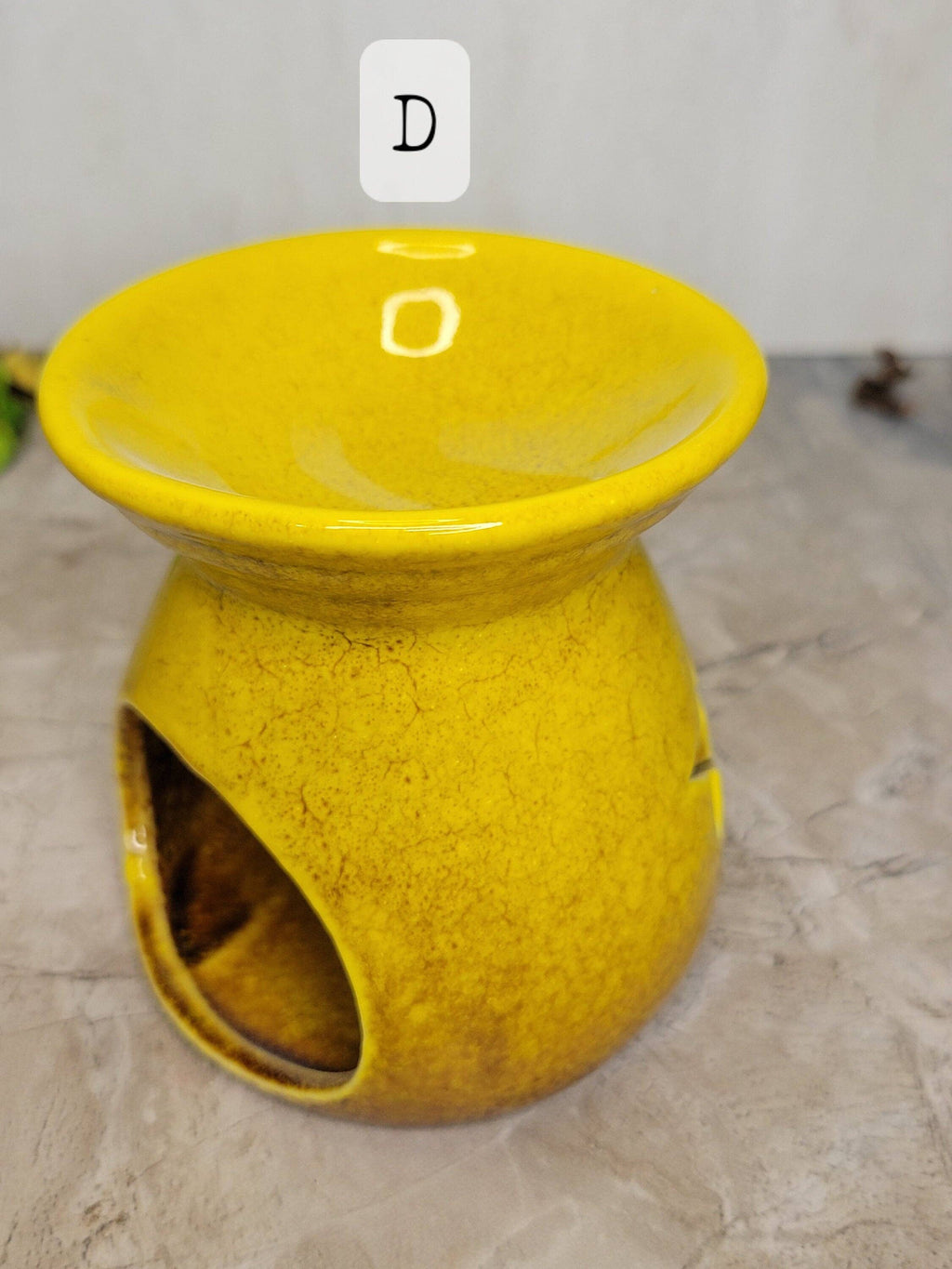 A bright yellow ceramic oil burner lamp with a textured glaze, featuring a wide top bowl, a side cutout for a candle holder, and a small white label with the letter 'D' near the rim, set on a gray stone surface.