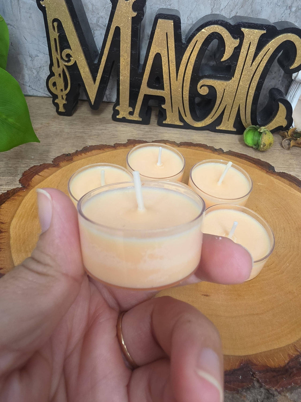 A hand holds a small clear glass tealight candle with pale peach wax in front of a wooden slice tray holding four more matching candles. A decorative black-and-gold 'MAGIC' sign and green leaves appear in the background.