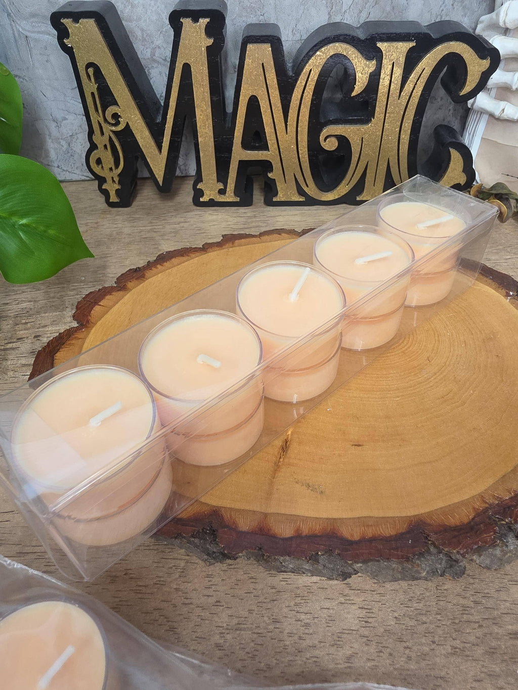 A set of six small tea light candles in a clear plastic tray, displayed on a round wood slice with a decorative 'MAGIC' sign in the background.