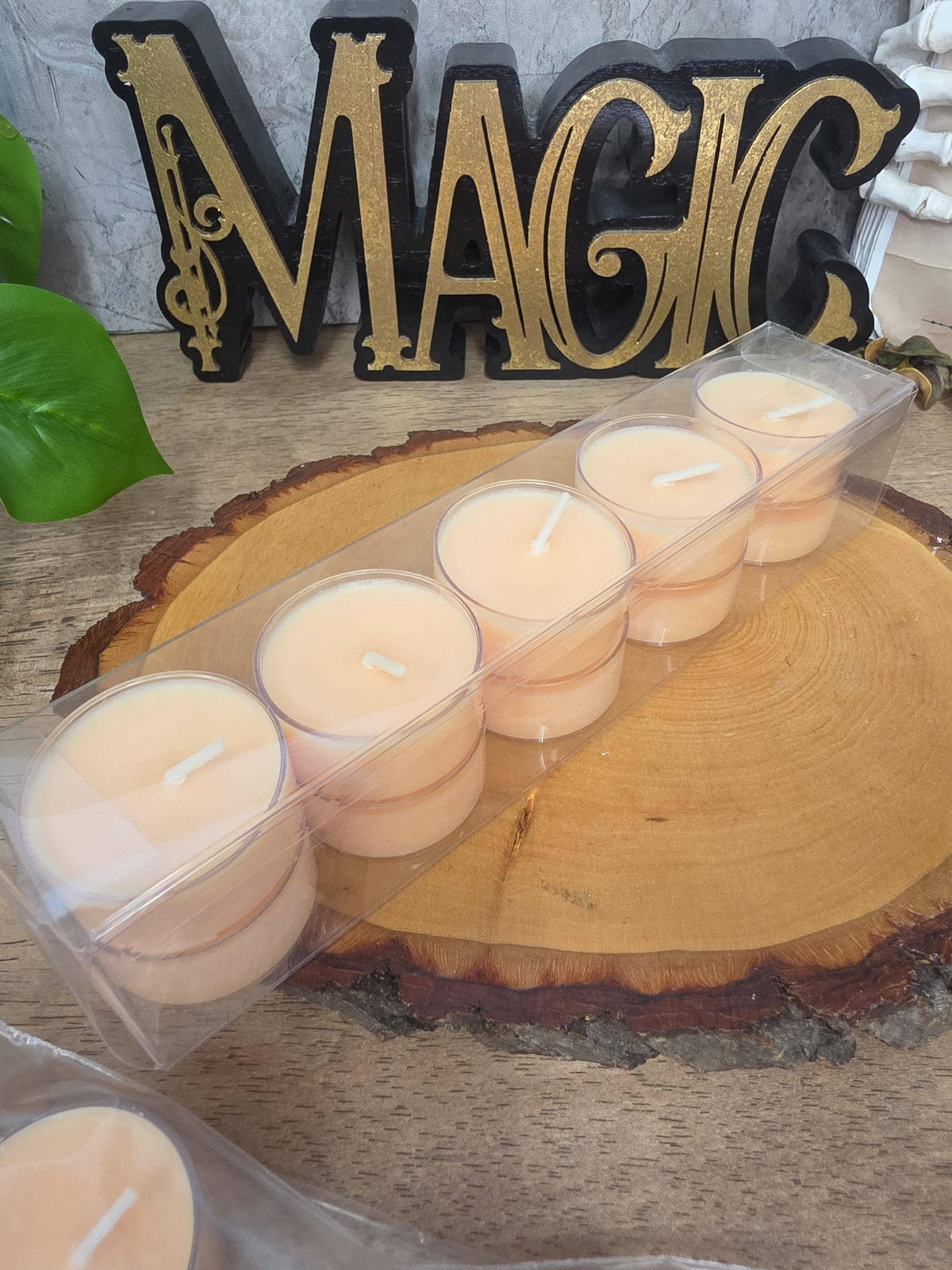 A set of six small tea light candles in a clear plastic tray, displayed on a round wood slice with a decorative 'MAGIC' sign in the background.