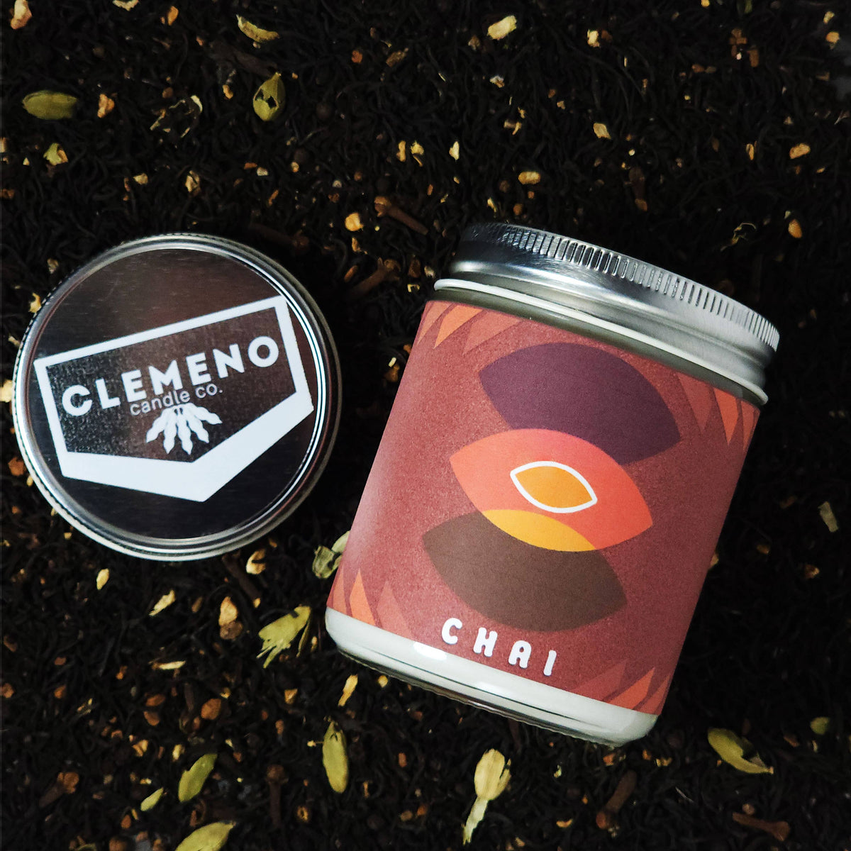 A small glass candle labeled 'CHAI' by Clemeno Candle Co., shown next to its lid on a dark tea-leaf background.