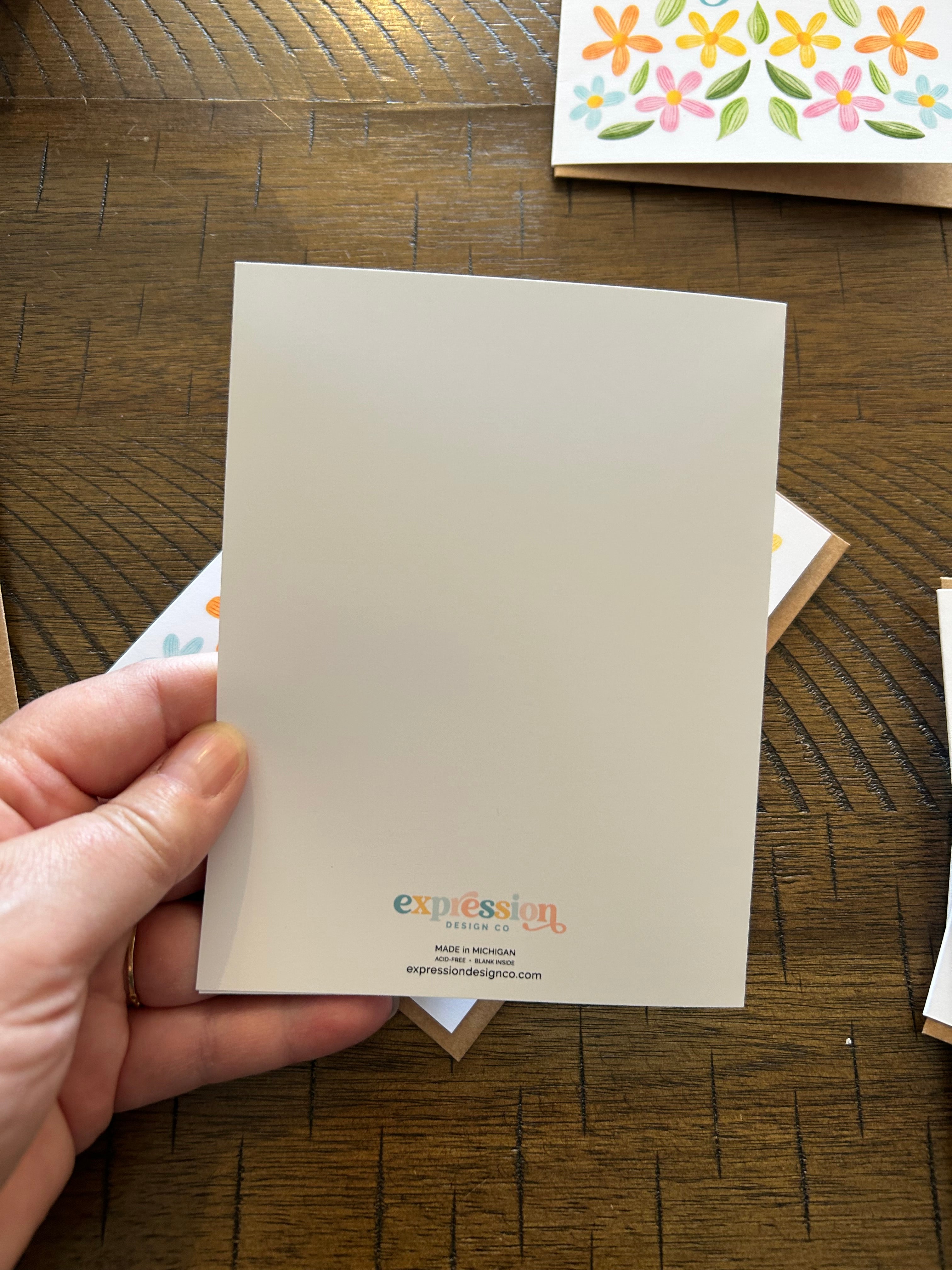 Hand holding a greeting card showing back of the card with a floral card in the background on a wooden surface