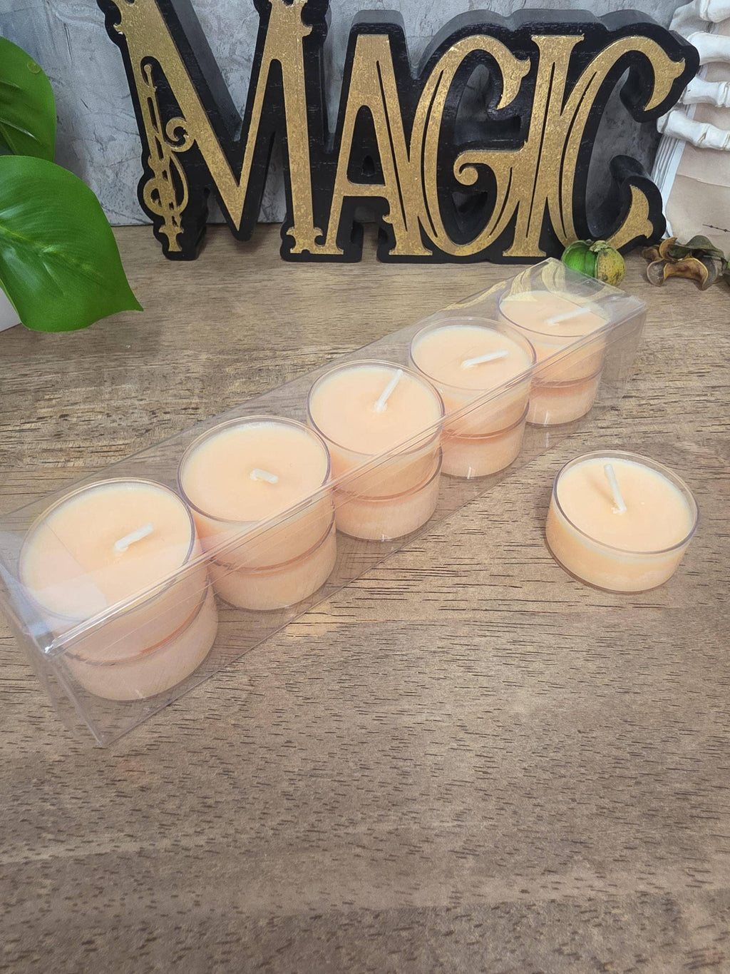 A set of small tea light candles in a clear plastic tray, with one candle placed beside the tray on a wooden surface. A decorative gold and black 'MAGIC' sign and green leaves are visible in the background.
