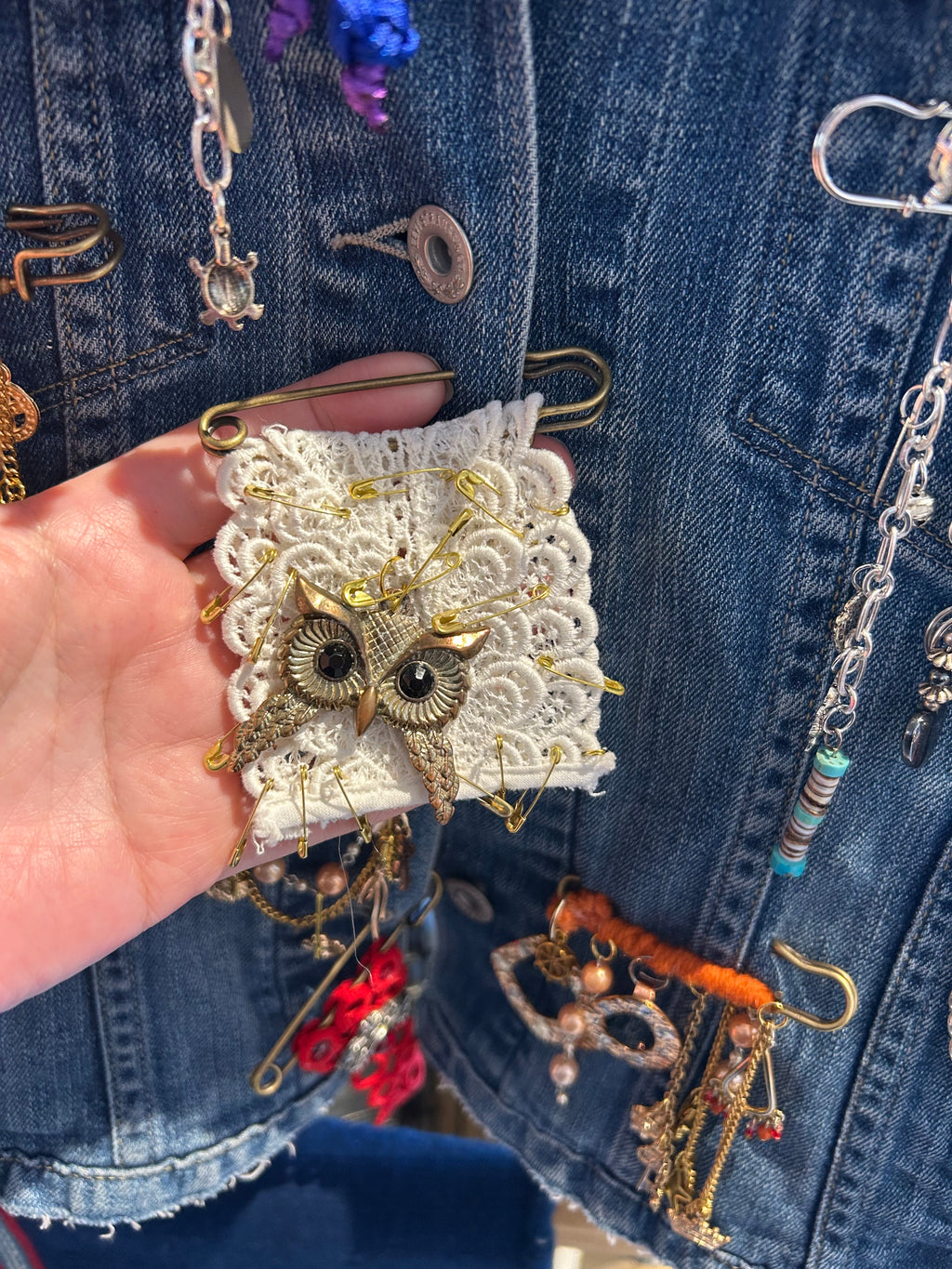 Hand holding a decorative owl patch over a denim background with various jewelry items.