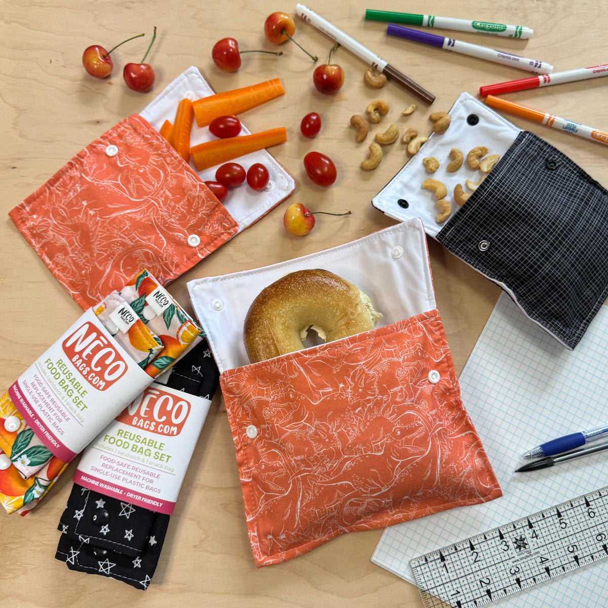 A flat-lay of reusable fabric pouches and a matching food bag set on a light wood surface, surrounded by school and kitchen props including cherries, carrots, nuts, markers, a bagel, a notebook, and pens. The pouches are shown in orange, white, and black patterns.