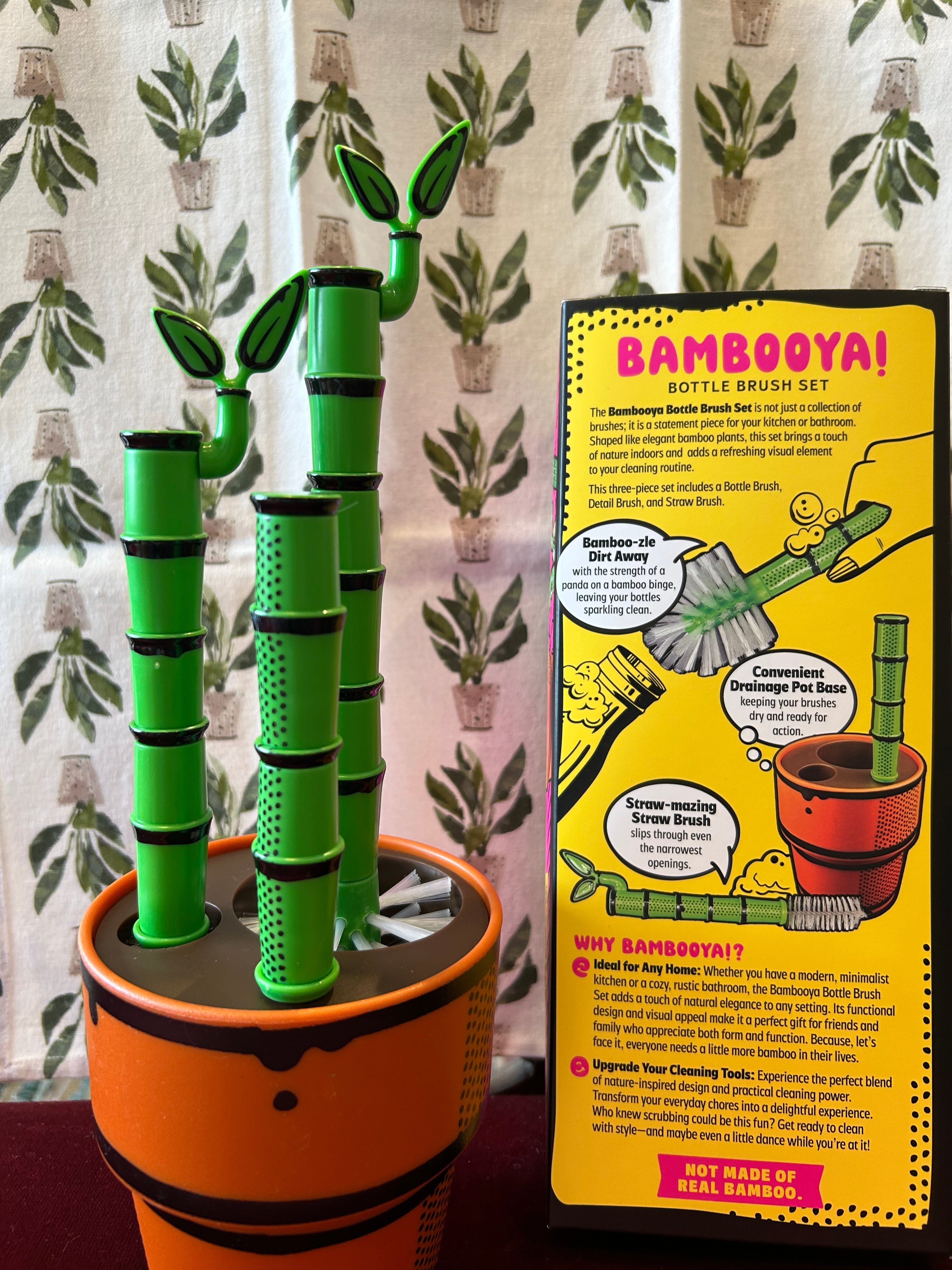 Green Bamboo Looking Bottle Brush Set with orange plant pot looking base with a Peace Lily Tea Towel by Mahogany, featuring a colorful box in the background.