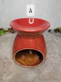 A ceramic wax warmer with a glossy terracotta red finish, featuring a wide shallow saucer top and an open base. A white label with the letter 'A' is overlaid near the top.