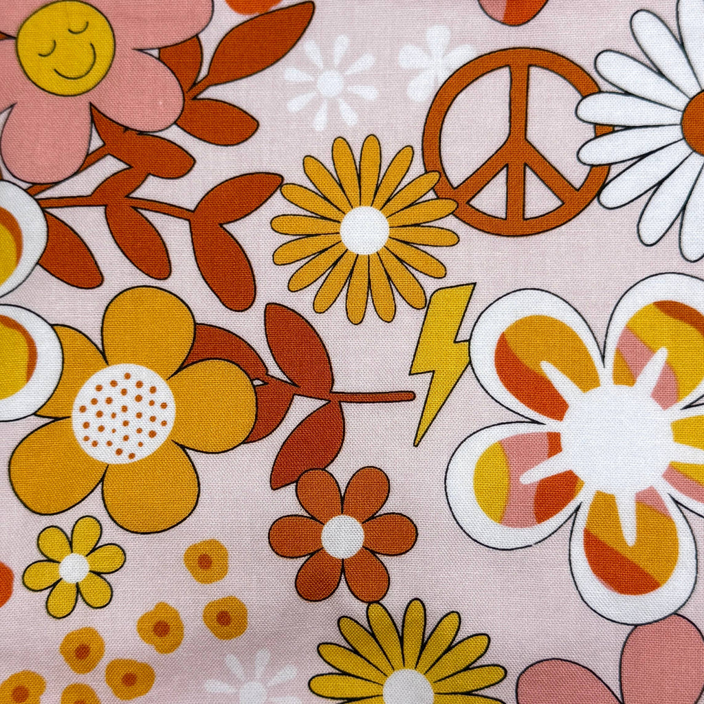 A close-up of a retro 1970s floral fabric print featuring yellow, orange, and pink flowers, peace sign, lightning bolt, and daisies on a dusty pink background.