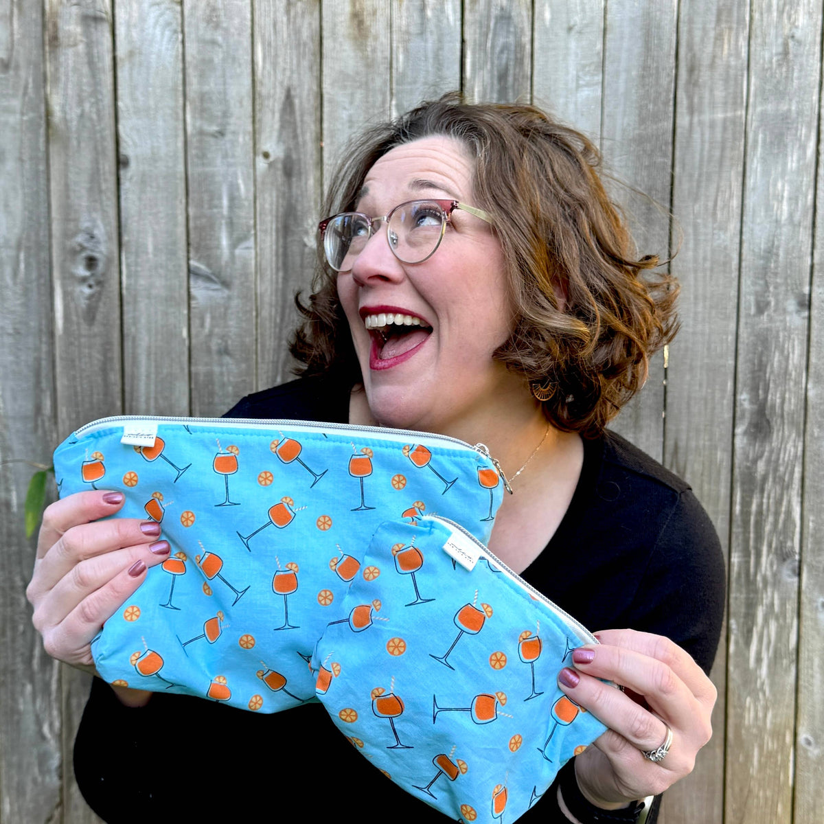 A woman with glasses and brown hair is smiling and holding a light blue zippered pouch decorated with an orange cocktail glass pattern, set against a wooden fence background.