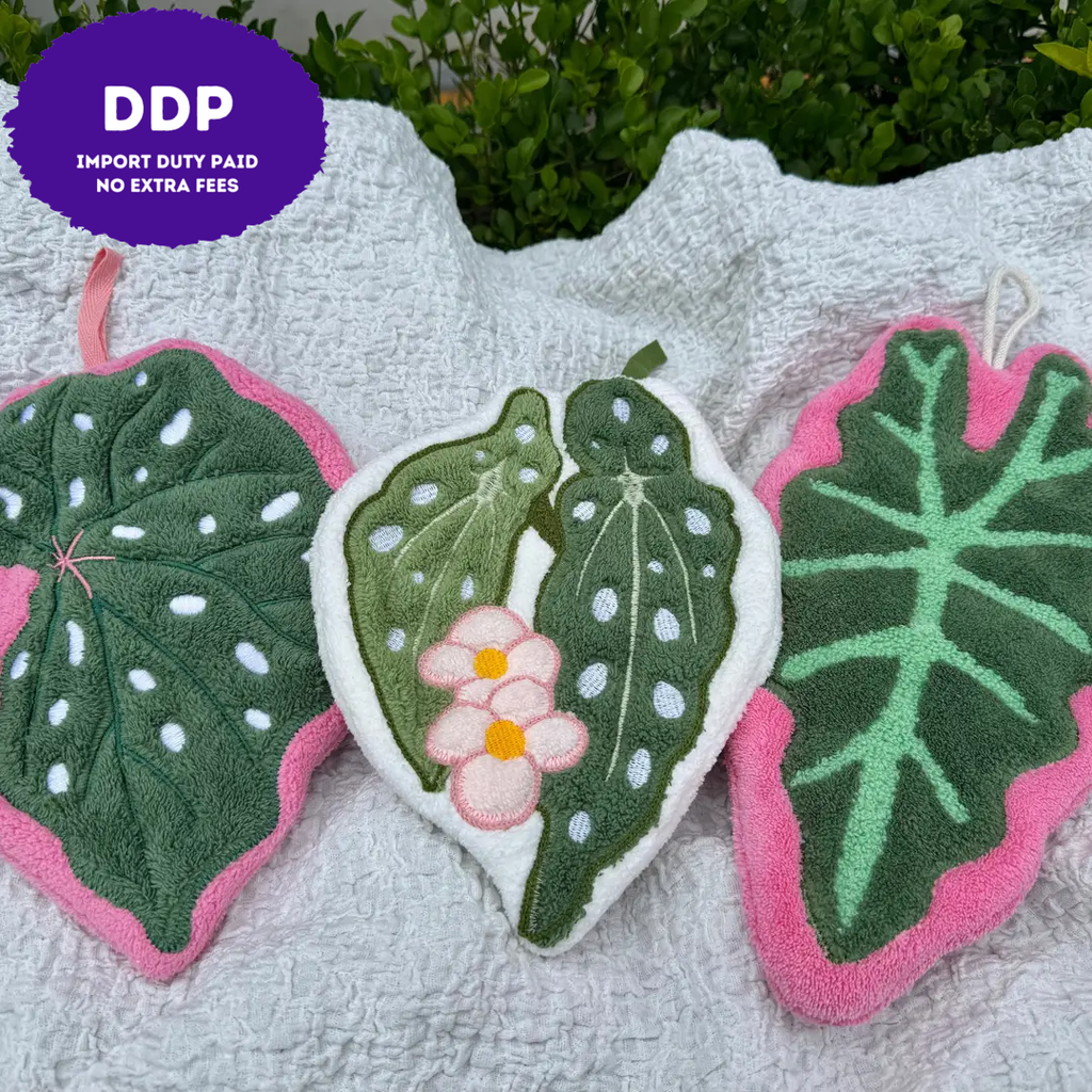 Three plush hand towels shaped like tropical leaves are arranged on a white textured blanket outdoors with green foliage in the background. The towels have dark green centers, pink edges, and white polka dots, with one towel featuring a small pink flower motif.