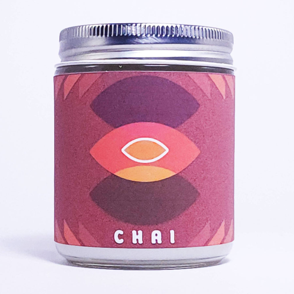 A glass jar candle with a silver metal lid and a textured pinkish-red label featuring a stylized eye design and the word 'CHAI' at the bottom, set against a plain white background.
