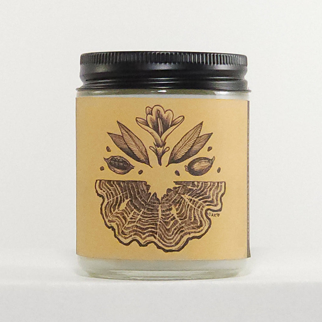 A clear glass jar candle with a black metal screw-top lid and a tan paper label featuring a dark brown botanical illustration of a flower and leaves above a wing-like pattern. The candle is photographed against a plain white background.