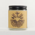 A clear glass jar candle with a black metal screw-top lid and a tan paper label featuring a dark brown botanical illustration of a flower and leaves above a wing-like pattern. The candle is photographed against a plain white background.