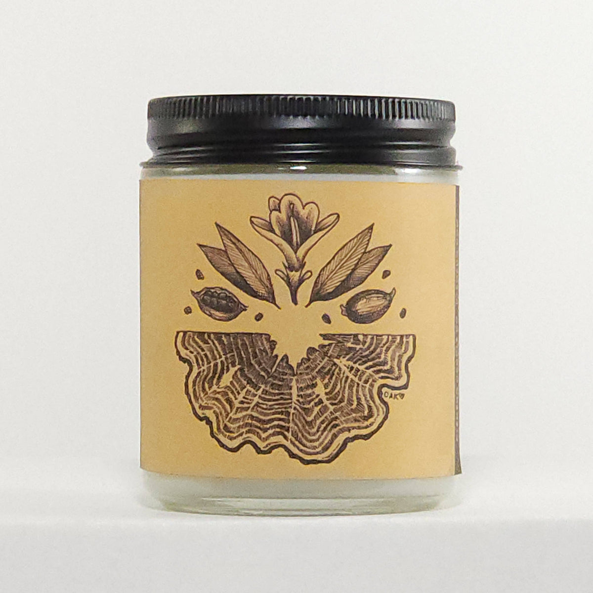 A clear glass jar candle with a black metal screw-top lid and a tan paper label featuring a dark brown botanical illustration of a flower and leaves above a wing-like pattern. The candle is photographed against a plain white background.