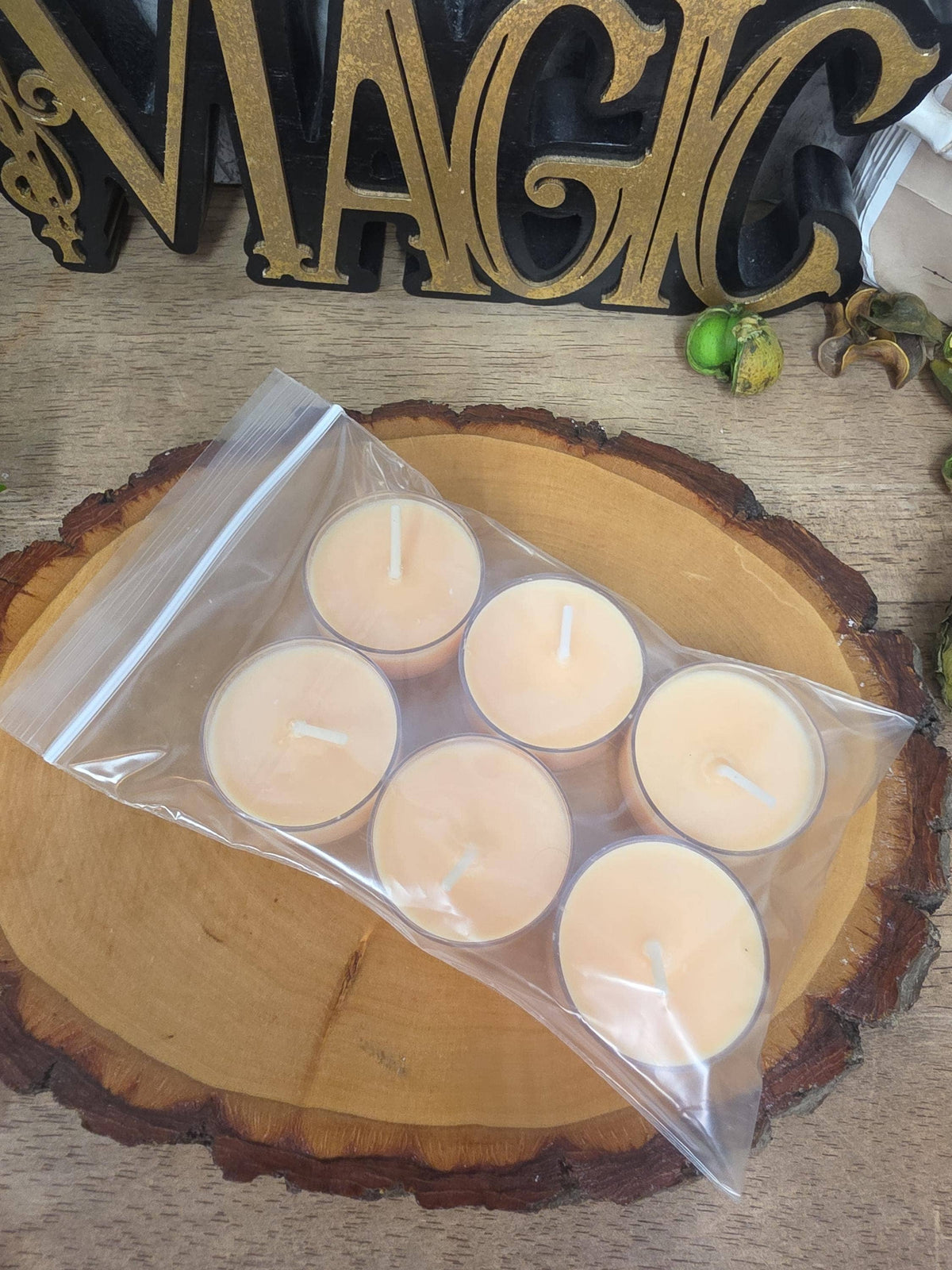 Six small tea light candles in a clear plastic bag, placed on a round wood slice coaster. A decorative gold and black 'MAGIC' sign is visible in the background on a wooden surface.