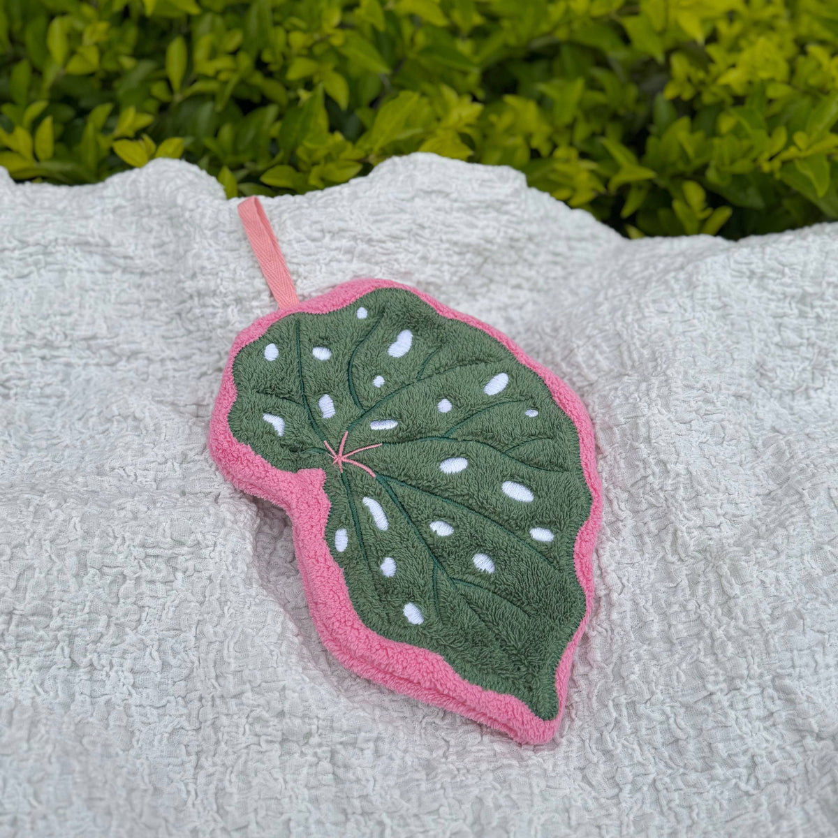 A leaf-shaped hand towel with a green center, white spots, and a pink border, resting on a textured white towel with green foliage in the background.