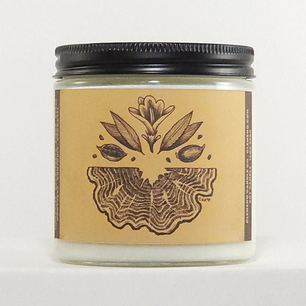 A clear glass jar containing a white wax candle, sealed with a black metal lid and wrapped in a tan paper label featuring a dark brown botanical illustration. The brand name 'SACV' is visible in small letters on the right side of the label.