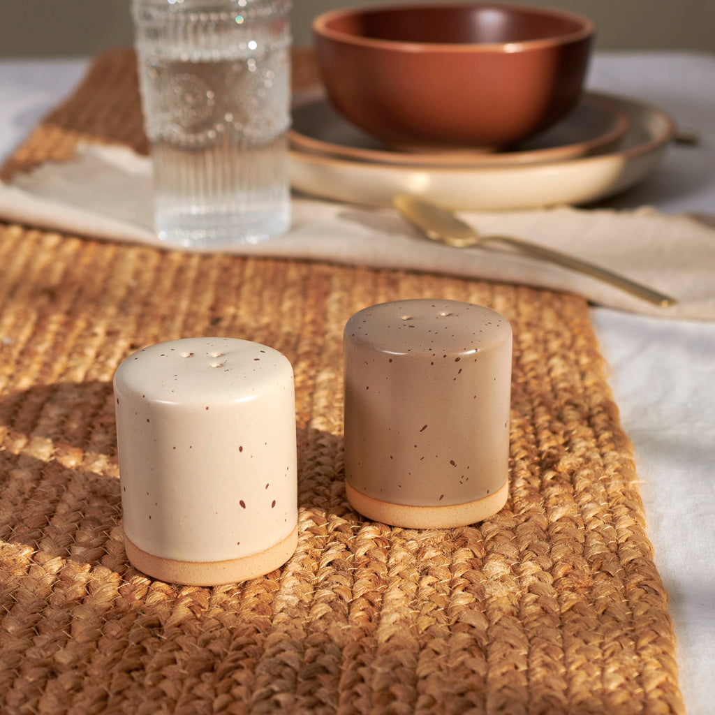 Two ceramic salt and pepper shakers on a woven placemat with a glass of water and bowl in the background.
