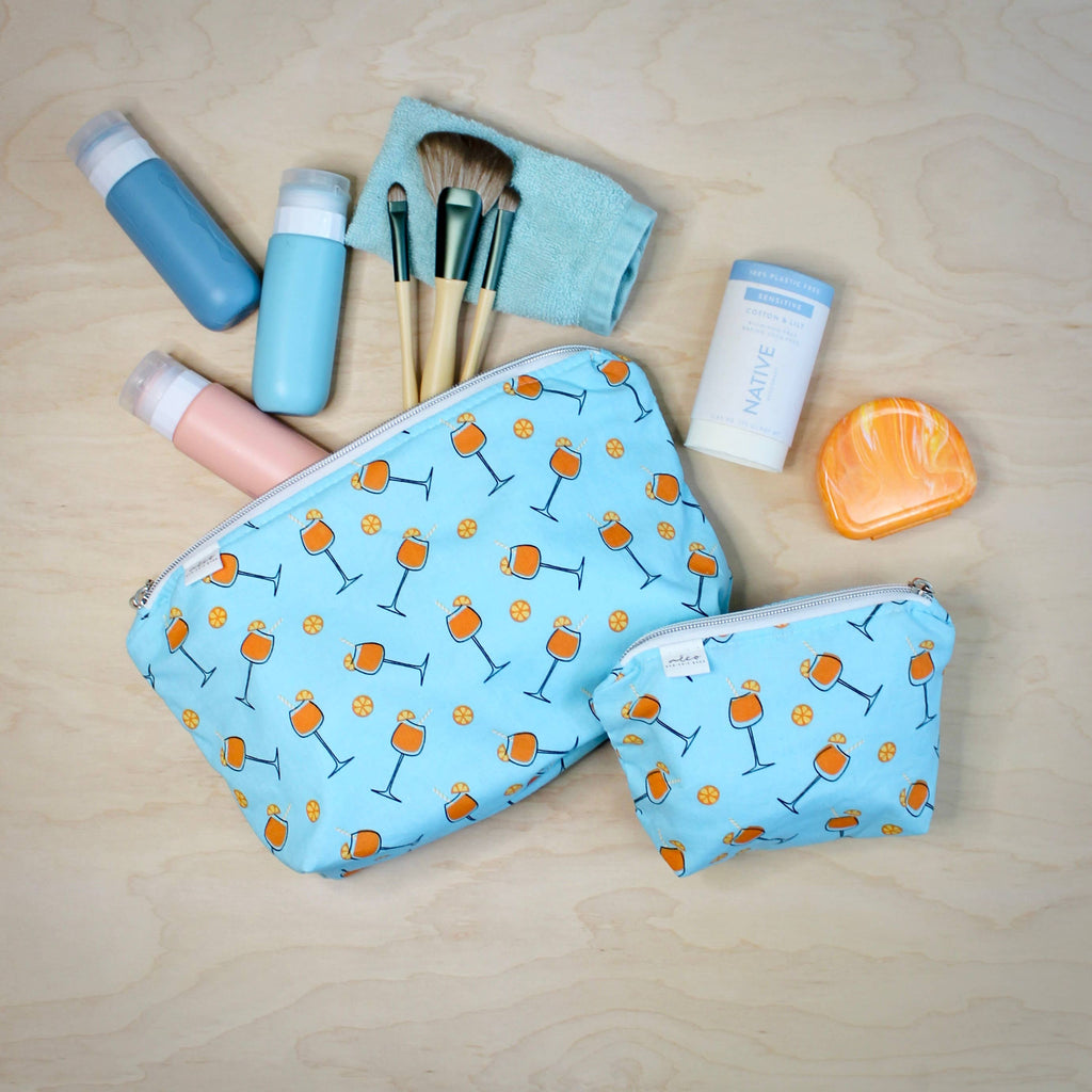 A flat-lay of two light blue zippered cosmetic pouches with an orange cocktail glass and yellow flower pattern, surrounded by travel-sized toiletries on a light wood background.