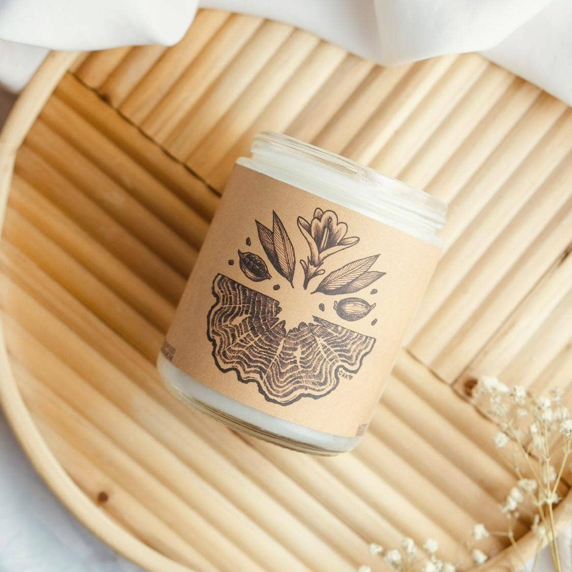 A glass jar candle with a kraft paper label featuring a black botanical illustration, styled on a round bamboo tray with dried white flowers.