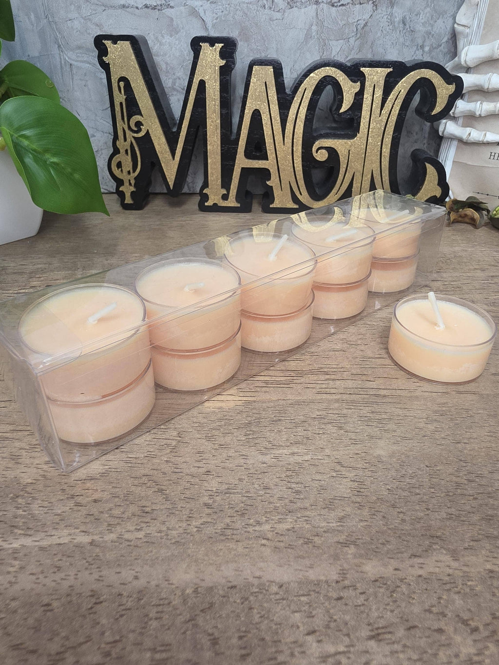A set of small tea light candles in a clear plastic tray, with one candle placed beside it on a wooden surface. A decorative black and gold 'MAGIC' sign and a potted plant are visible in the background.