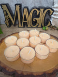 Nine small tea light candles with pale peach wax are arranged on a round wood slice tray, with a large decorative 'MAGIC' sign in the background.