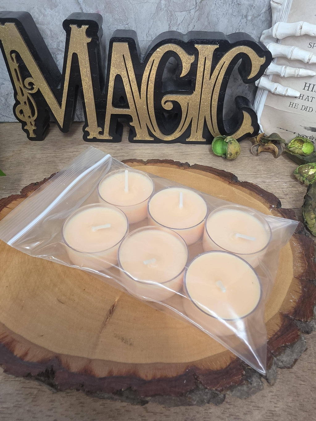 Six small tea light candles in a clear resealable bag, placed on a round wood slice. A decorative black-and-gold 'MAGIC' sign and other styled props appear in the background.