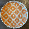 A round bamboo woven basket tray with a geometric pattern, predominantly beige and orange in color.